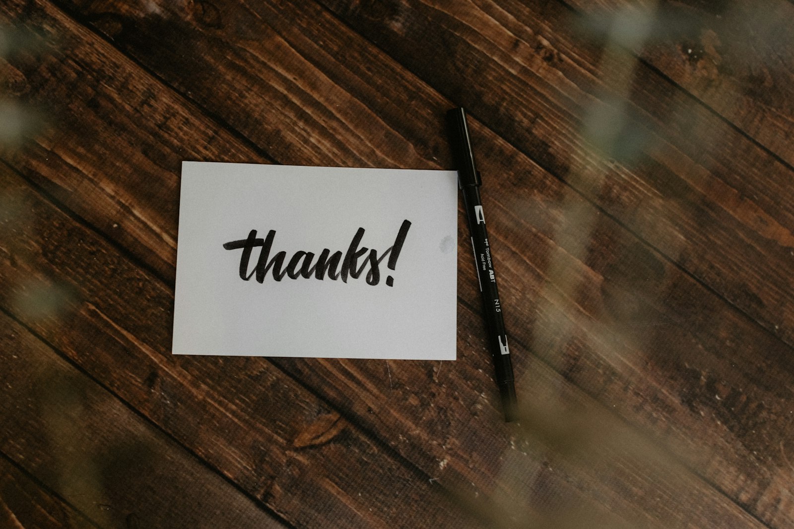 Small white card with the handwritten word thanks next to a black pen on a dark wooden table