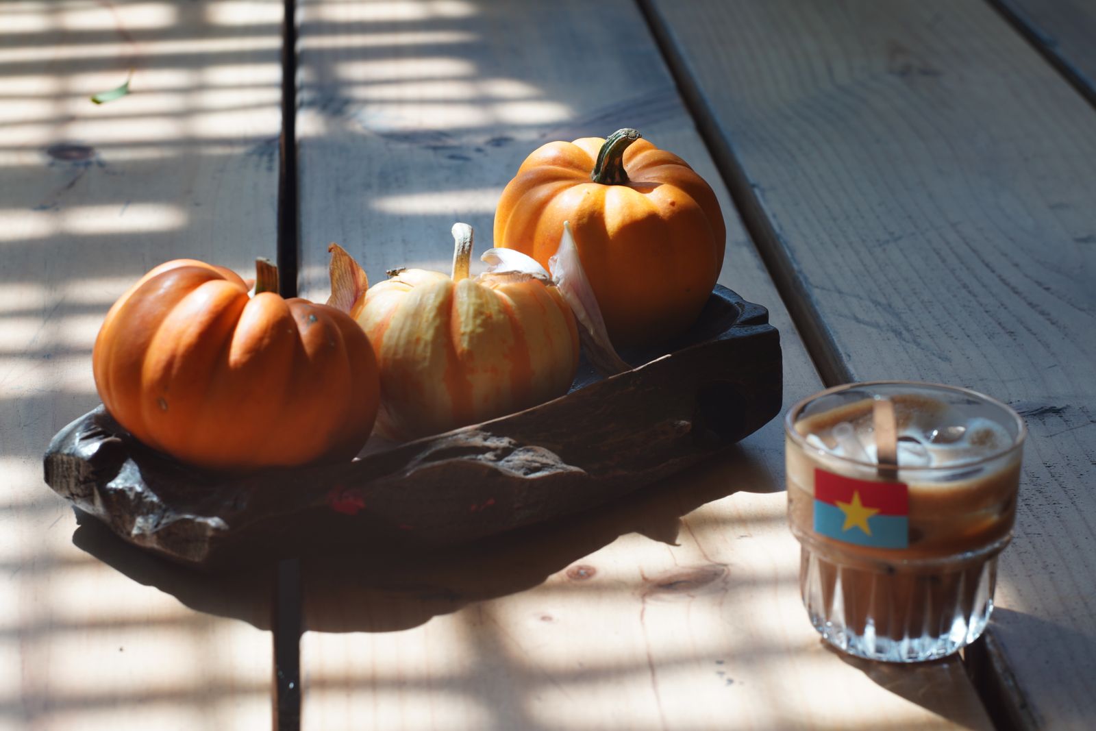 Small pumpkins on a dark tray beside a glass of iced coffee on a sunlit wooden table